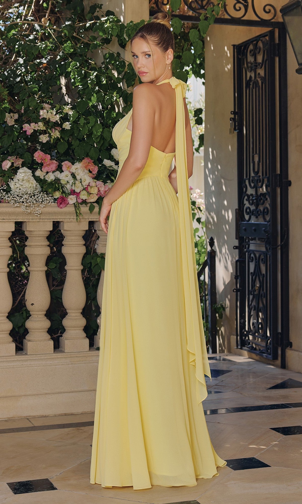 A woman wears the Ladivine T715 Long Chiffon Halter Prom Dress on a tiled patio, facing away, with greenery and flowers in the background.